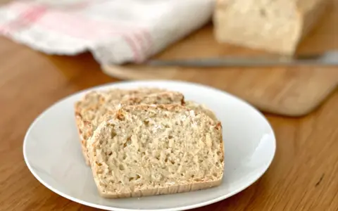 Skivor av hembakat bröd på en vit tallrik, perfekt för snabb bakning. En bit bröd ligger på en skärbräda i bakgrunden.
