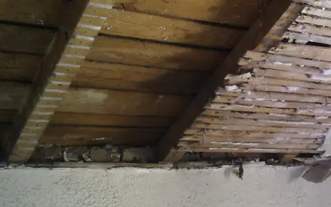 Vue intérieure d'une charpente en bois, montrant des lattes et des planches, avec des traces de plâtre. Un pare-pluie par l'intérieur semble avoir été appliqué.