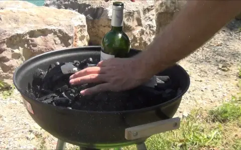 Main d'homme plaçant une bouteille de vin verte sur du charbon de bois dans un barbecue, étape pour comment allumer un barbecue.