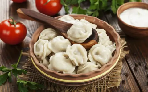 Pielmieni co to? Pyszne, gotowane pierożki w glinianej misce, z drewnianą łyżką, pomidorami i śmietaną w tle.