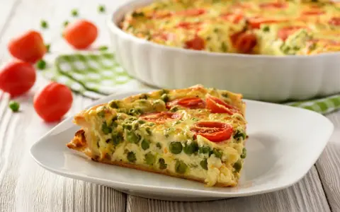 Ein Stück vegetarische Quiche mit Erbsen und Tomaten auf einem Teller. Das ganze Gericht ist im Hintergrund zu sehen.