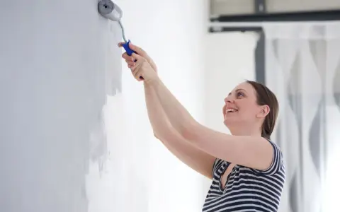 Une femme souriante apprend comment peindre un mur avec un rouleau. Elle applique une couche de peinture grise sur un mur blanc.
