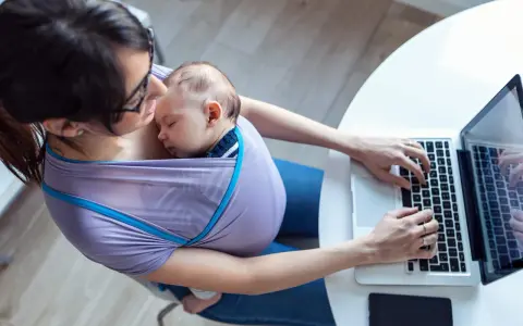 Kobieta w chuście nosi śpiące dziecko i pracuje na laptopie. Czy na macierzyńskim można pracować? Tak, to dowód.