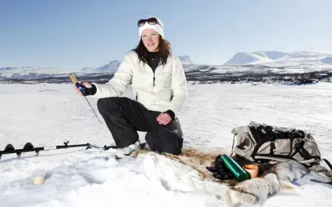 Kvinna ispirar isfiske i Kiruna. Hon har fiskeutrustning och en ryggsäck bredvid sig.
