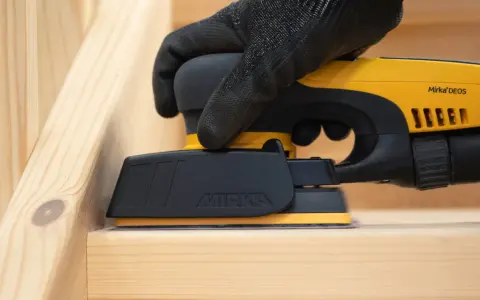 En hand med handske slipar en träyta med en Mirka DEOS-slipmaskin. Perfekt för att slipa borr och andra trädetaljer.