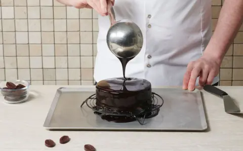 Un chef verse un nappage chocolat brillant sur un gâteau. Des pépites de chocolat sont à côté.