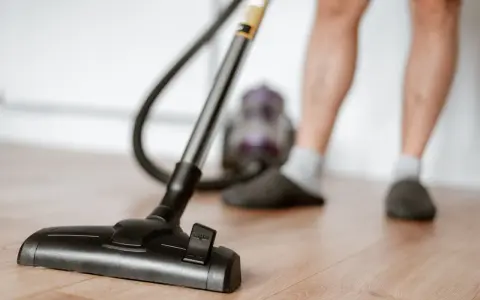 Odkurzacz na drewnianej podłodze, część nóg w kapciach i skarpetkach. To jeden z obowiązków domowych po angielsku – "vacuuming".