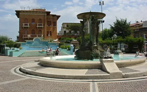 Fontana con statue e tartarughe in una piazza di Cattolica centro, con edifici storici e persone che si godono il sole.