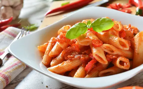 Eine Schüssel Penne Arrabiata, garniert mit Basilikum, mit scharfen Chilis und Knoblauch im Hintergrund.