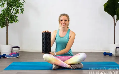 Frau zeigt, wie benutzt man eine Faszienrolle für das Training. Sie sitzt auf einer blauen Matte mit Gewichten und Pflanzen im Hintergrund.