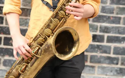 Hände spielen ein goldenes Saxophon. Obwohl es aus Metall ist, ist das Saxophon ein Holzblasinstrument, da der Ton durch ein Rohrblatt erzeugt wird.