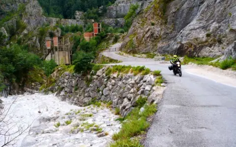 Un motociclista percorre una strada tortuosa tra le Alpi Apuane, con un fiume di sassi e ruderi industriali sullo sfondo.