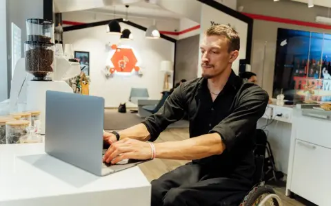 Mężczyzna na wózku inwalidzkim pracuje przy laptopie. To przykład integracji i pracy z grupą inwalidzką.