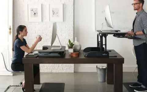 Un homme et une femme travaillent sur des bureaux réglables en hauteur. Une pause obligatoire pour le travail debout est essentielle pour leur bien-être.