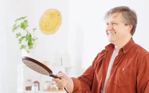Un homme souriant s'amuse à retourner une crêpe en plein vol avec une poêle.