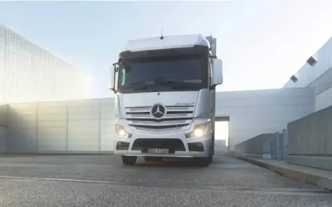 Biały Mercedes Actros MP4 z włączonymi światłami stoi przed nowoczesnym budynkiem.