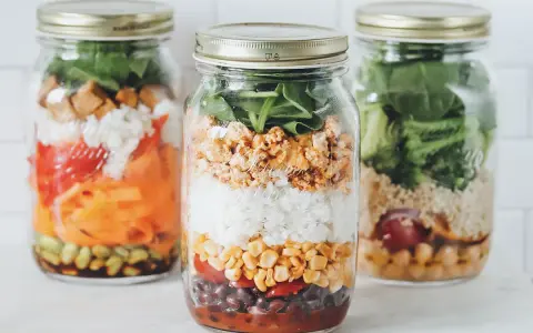 Drei bunte Schichtsalate im Glas, bereit für unterwegs. Jedes Glas ist ein kleines Kunstwerk mit Reis, Gemüse und Proteinen.