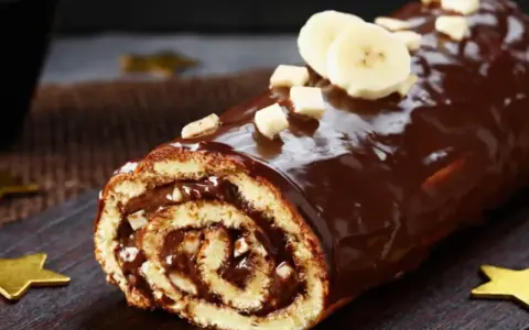 Bûche roulée au chocolat, garnie de crème et recouverte d'un glaçage chocolat beurre brillant, décorée de rondelles de banane et de noisettes concassées.