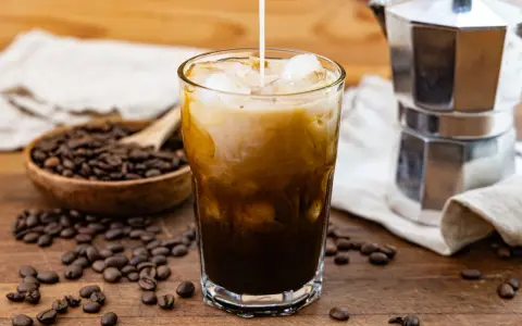 Milch fließt in ein Glas mit Eis und Kaffee. Die **cold brew funktion** wird durch die Kaffeebohnen und die Mokkakanne im Hintergrund unterstrichen.