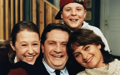 En glad familj, som hämtad ur svenska komedier, ler mot kameran. Pappa, mamma, dotter och son.