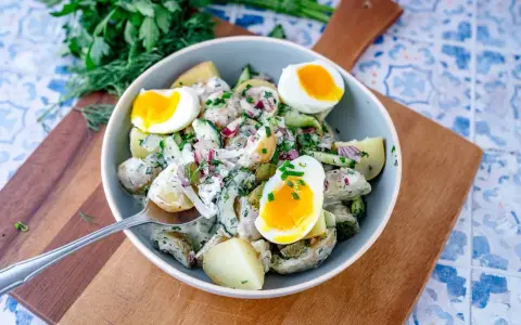 Schüssel mit cremigem Kartoffelsalat mit Gurken, Zwiebeln und gekochten Eiern, garniert mit frischen Kräutern.