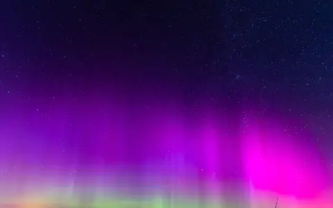 Nocne niebo rozświetlone fioletową i zieloną zorzą polarną, kiedy jest zorza polarna. Gwiazdy migoczą nad ciemnym horyzontem.
