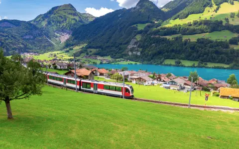 A red and white train travels along the **Golden Pass Line Lucerne to Interlaken**, passing a village by a turquoise lake, with lush green mountains in the background.