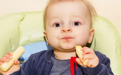 Mały chłopiec z radością zajada chrupki kukurydziane dla niemowląt, siedząc w krzesełku.
