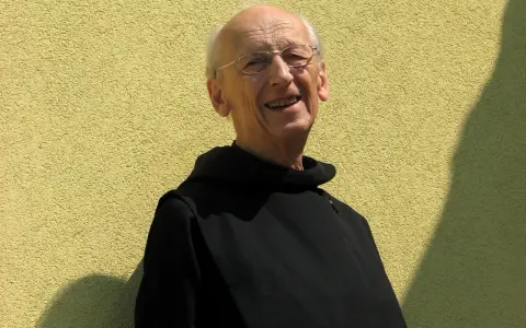 Uśmiechnięty starszy mężczyzna w czarnym habicie, z okularami na nosie, wygląda jak pobożna dewota.