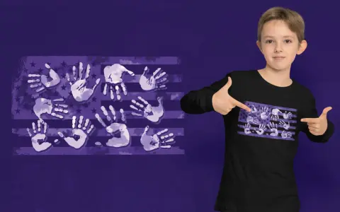Un niño señala una camiseta negra con un diseño de bandera americana hecho de huellas de manos moradas. ¡Un diseño creativo para pintar camisetas con las manos!