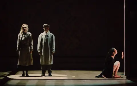 Magdalena Twardowska w scenie teatralnej. Dwie postacie w płaszczach stoją w świetle, obok nich kobieta w czarnej sukni klęczy przy pionowym świetle.