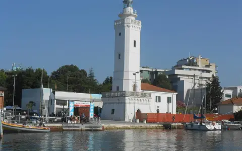 Il faro di Rimini, con la sua torre bianca e il porto con barche a vela, si staglia contro il cielo azzurro.