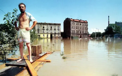 Mężczyzna na prowizorycznej tratwie w zalanej wodą ulicy. Budynki i samochody toną w brązowej wodzie. To obraz powodzi 1997.