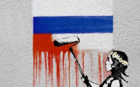 Dziecko w wianku maluje wałkiem czerwoną farbę na fladze Rosji.