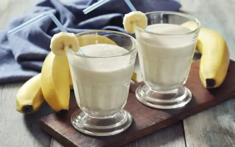 Dwa bananowe koktajle proteinowe w szklankach z rurkami i plasterkami banana. Idealne, gdy pić shake proteinowy po treningu.