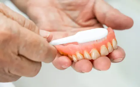 Dłonie czyszczą protezę zębową szczoteczką, pokazując jak usunąć kamień z plastiku.