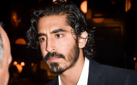 Primer plano de un hombre con cabello oscuro y rizado, y una barba poco poblada larga. Viste una camisa blanca y un traje oscuro.