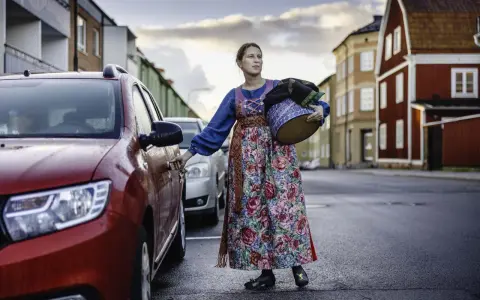 Lina Odell, klädd i folkdräkt, öppnar bildörren med en korg i famnen.