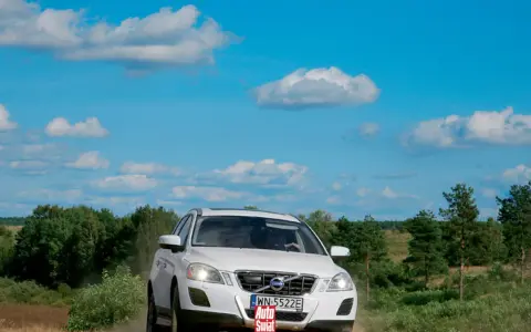 Białe Volvo XC60 T6 jedzie po piaszczystej drodze wśród zieleni, pod błękitnym niebem z chmurami.