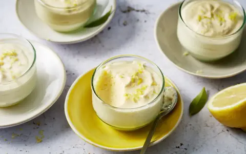 Plusieurs verrines de mousse au citron, garnies de zestes, sur des assiettes claires et jaunes. Un citron coupé est visible à droite.