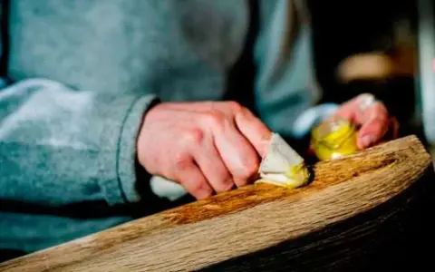 Manos aplicando cera a un trozo de madera, restaurando su brillo. El proceso de decapar madera revela su belleza natural.