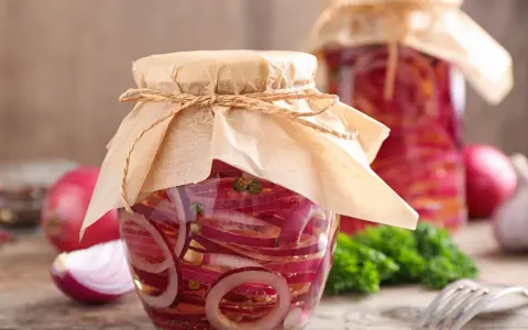 Zwei Gläser mit eingelegten Zwiebeln, verziert mit Papier und Schnur. Daneben liegen frische Zwiebeln und ein Gabel.