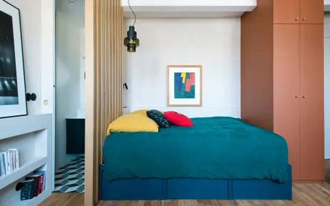 Une chambre cabine cosy avec un lit bleu canard, des coussins colorés et une armoire ocre. Un paravent en bois sépare l'espace nuit de la salle de bain.