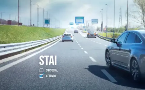 Sulla strada, auto e camion procedono. "STAI ATTENTO" è scritto, ricordando l'art. 79 CDS.