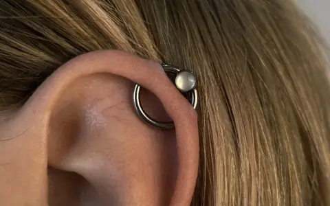 Oryginalny kolczyk orbital piercing z białym kamieniem na uchu, otoczony blond włosami.