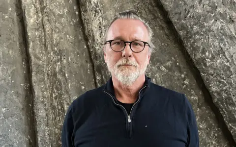 Thomas Olsson, en man med glasögon och grått skägg, står framför en grov, stenliknande vägg.