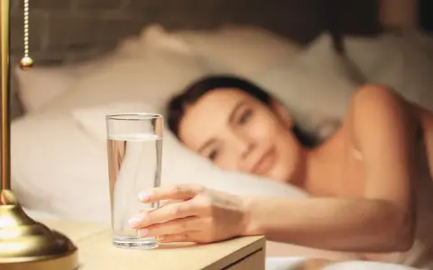 Une main attrape un verre d'eau posé à droite sur une table de chevet, une femme se réveille en arrière-plan.