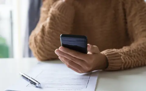 Persona revisa su teléfono móvil, modelo 035, mientras tiene un bolígrafo y un formulario de solicitud sobre la mesa.