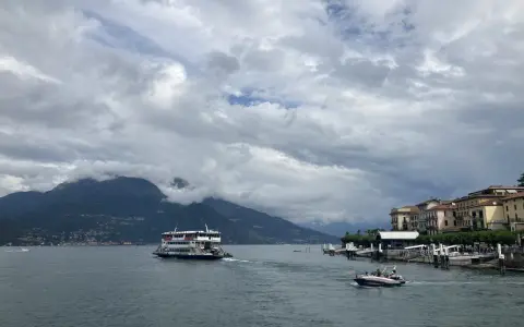 Promenada nad jeziorem Como, z górami w tle i statkiem wycieczkowym. W oddali widać urokliwe miasteczko.