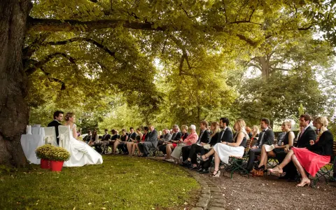 Hochzeit im Freien unter einem Baum. Gäste sitzen auf Bänken. Tolle Ideen zur Hochzeit für eine romantische Zeremonie.
